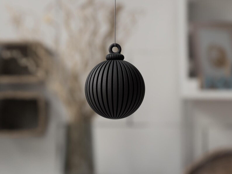 May include: A black, spherical ornament with vertical grooves, suspended by a thin string. The ornament has a decorative top with a loop for hanging. The background is blurred, suggesting a home decor setting.