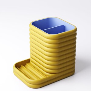 May include: A yellow desk organizer with a blue interior and a small tray. The organizer has a ribbed design and is made of a solid material. The tray has a ribbed design. The organizer is on a white background.