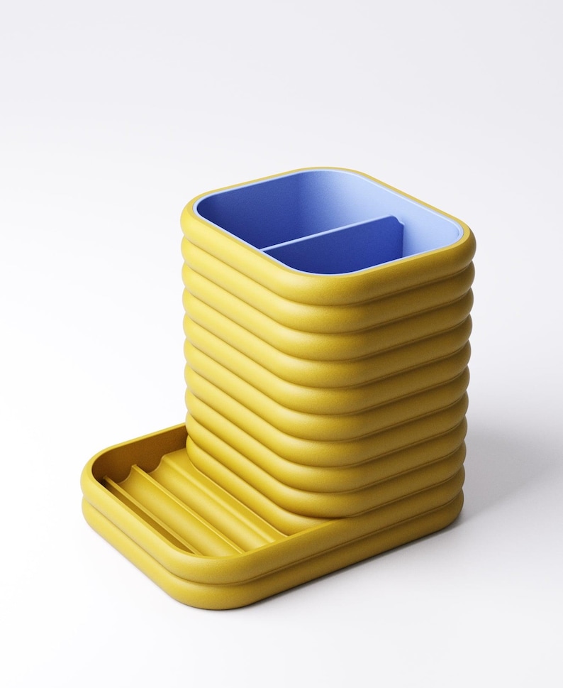 May include: A yellow desk organizer with a blue interior and a small tray. The organizer has a ribbed design and is made of a solid material. The tray has a ribbed design. The organizer is on a white background.