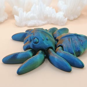 Op de afbeelding: Een blauwgroene iriserende spinnensculptuur met acht poten. De sculptuur heeft een textuur en staat tegen een lichte achtergrond met witte kristalformaties.