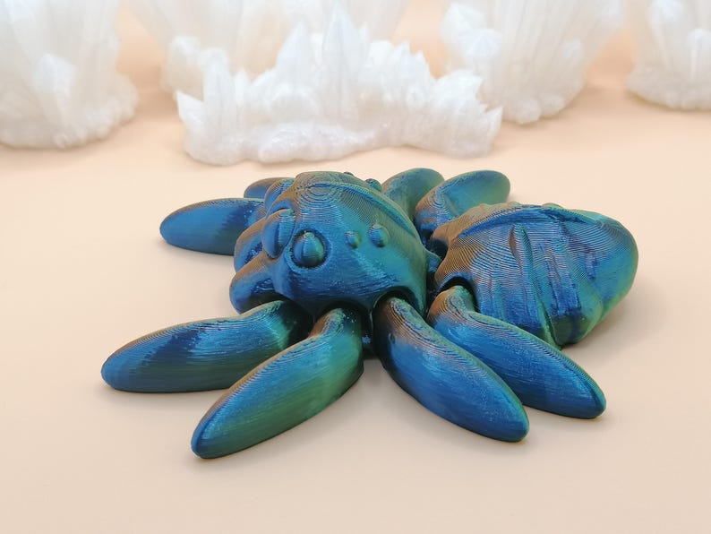 Op de afbeelding: Een blauwgroene iriserende spinnensculptuur met acht poten. De sculptuur heeft een textuur en staat tegen een lichte achtergrond met witte kristalformaties.