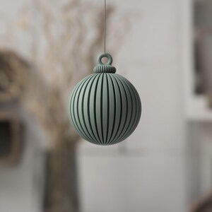 May include: A sage green, spherical ornament with vertical grooves and a loop for hanging. The decorative ball is suspended against a blurred background, suggesting a holiday or seasonal theme. The design is simple and modern.