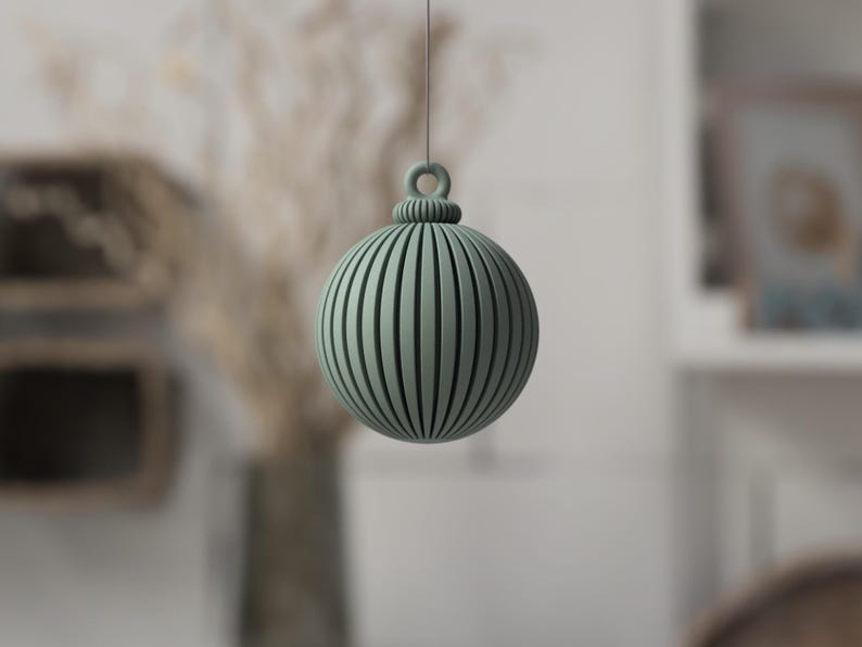 May include: A sage green, spherical ornament with vertical grooves and a loop for hanging. The decorative ball is suspended against a blurred background, suggesting a holiday or seasonal theme. The design is simple and modern.