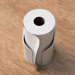 Op de afbeelding: Een witte papieren roldispenser met een rol wit papier. De dispenser is lichtgrijs van kleur en heeft een geribbeld ontwerp. Het papier is gedeeltelijk afgerold, wat de textuur van het papier laat zien. De dispenser staat op een houten oppervlak.