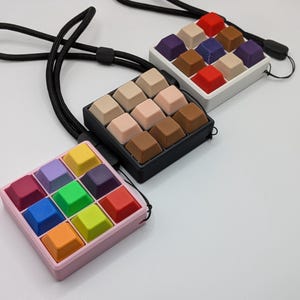 May include: Three square keycap pendants with black lanyards. One pendant is pink with colorful keycaps, another is black with neutral keycaps, and the last is white with a mix of colorful keycaps. The keycaps are arranged in a 3x3 grid.