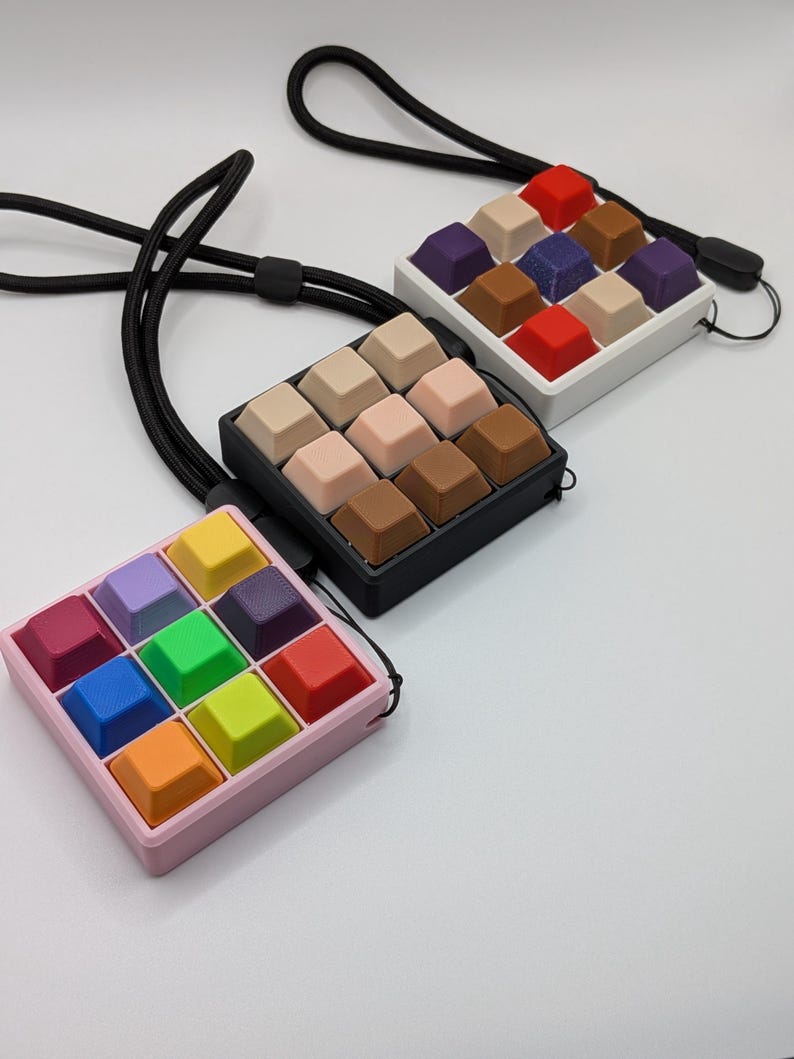 May include: Three square keycap pendants with black lanyards. One pendant is pink with colorful keycaps, another is black with neutral keycaps, and the last is white with a mix of colorful keycaps. The keycaps are arranged in a 3x3 grid.