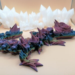 May include: An assortment of 3D-printed decorative items. The foreground features a series of rose and crystal-like structures with a gradient of colors, including blue, purple, and green. In the background, there are several white crystal-shaped objects.