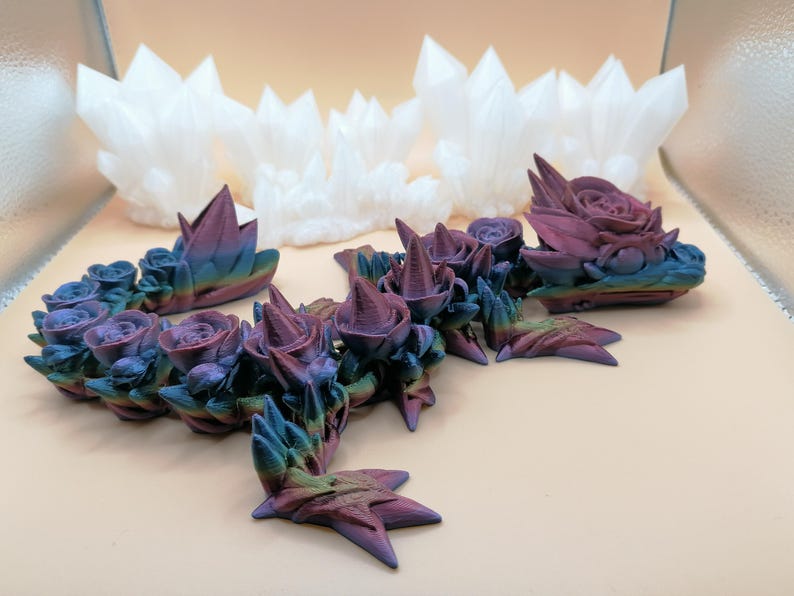 May include: An assortment of 3D-printed decorative items. The foreground features a series of rose and crystal-like structures with a gradient of colors, including blue, purple, and green. In the background, there are several white crystal-shaped objects.