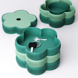 May include: A set of stackable flower-shaped storage containers in shades of green. The containers feature a ribbed exterior texture and a smooth interior. One container is open, holding a silver key with a black fob, while another lid sits nearby.