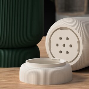 May include: A close-up of three ceramic objects. A dark green, ribbed, cylindrical container is in the background. In the foreground, a light beige, ribbed, cylindrical object with a perforated base and a matching small, ribbed, circular lid.