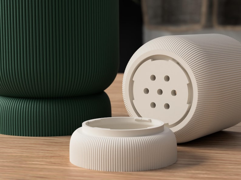 May include: A close-up of three ceramic objects. A dark green, ribbed, cylindrical container is in the background. In the foreground, a light beige, ribbed, cylindrical object with a perforated base and a matching small, ribbed, circular lid.