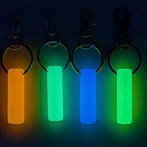 May include: Four keychains with glowing rectangular charms in orange, green, blue, and green. Each keychain has a silver-colored clasp and a ring. The background is dark, emphasizing the vibrant colors of the charms.