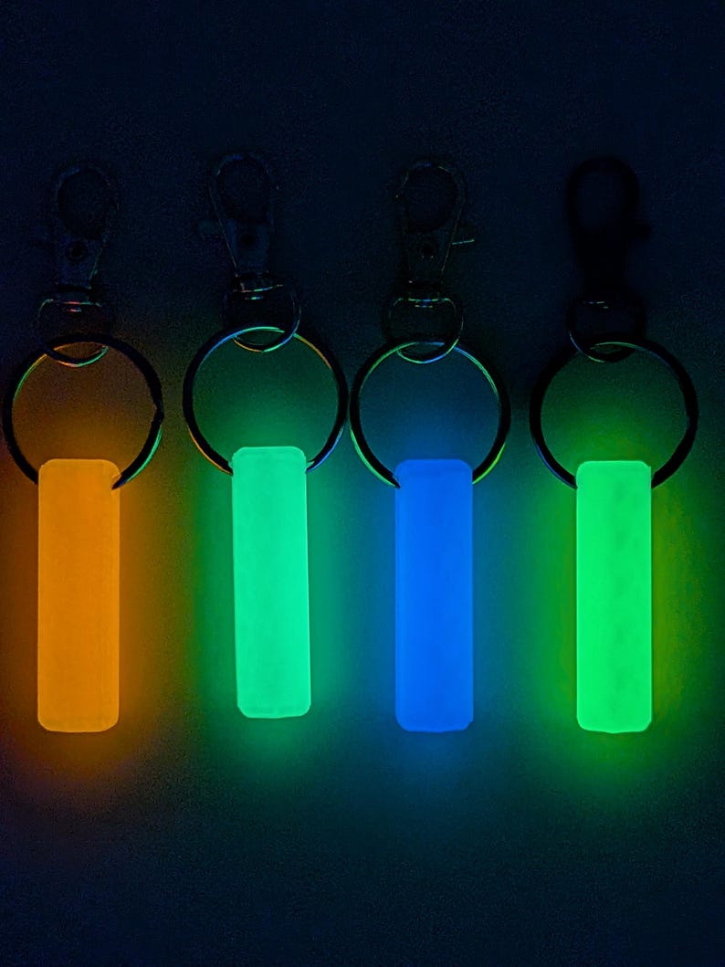 May include: Four keychains with glowing rectangular charms in orange, green, blue, and green. Each keychain has a silver-colored clasp and a ring. The background is dark, emphasizing the vibrant colors of the charms.