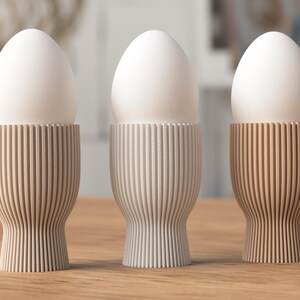 Pu&ograve; includere: Tre uova bianche poggiano in portauova decorativi. I portauova sono realizzati in un materiale di colore chiaro con scanalature verticali e ogni tazza ha una tonalit&agrave; beige unica. Le uova sono di un colore bianco brillante.