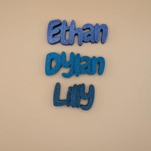 May include: Three blue glitter names: "Ethan", "Dylan", and "Lilly". The names are in a rounded font, each with a different shade of blue. The background is a plain, neutral beige colour.