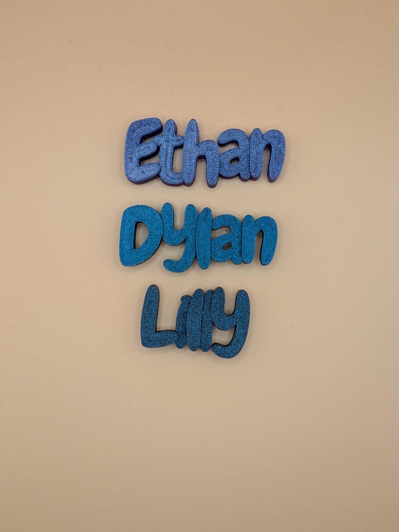 May include: Three blue glitter names: "Ethan", "Dylan", and "Lilly". The names are in a rounded font, each with a different shade of blue. The background is a plain, neutral beige colour.