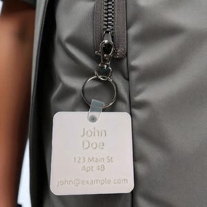 May include: A gray jacket with a silver zipper and a silver key ring attached. A white square tag with the name "John Doe", address, and email is attached to the key ring.