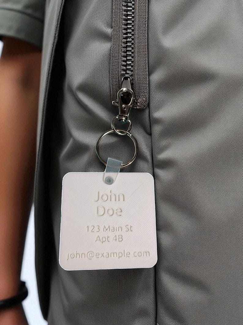 May include: A gray jacket with a silver zipper and a silver key ring attached. A white square tag with the name "John Doe", address, and email is attached to the key ring.