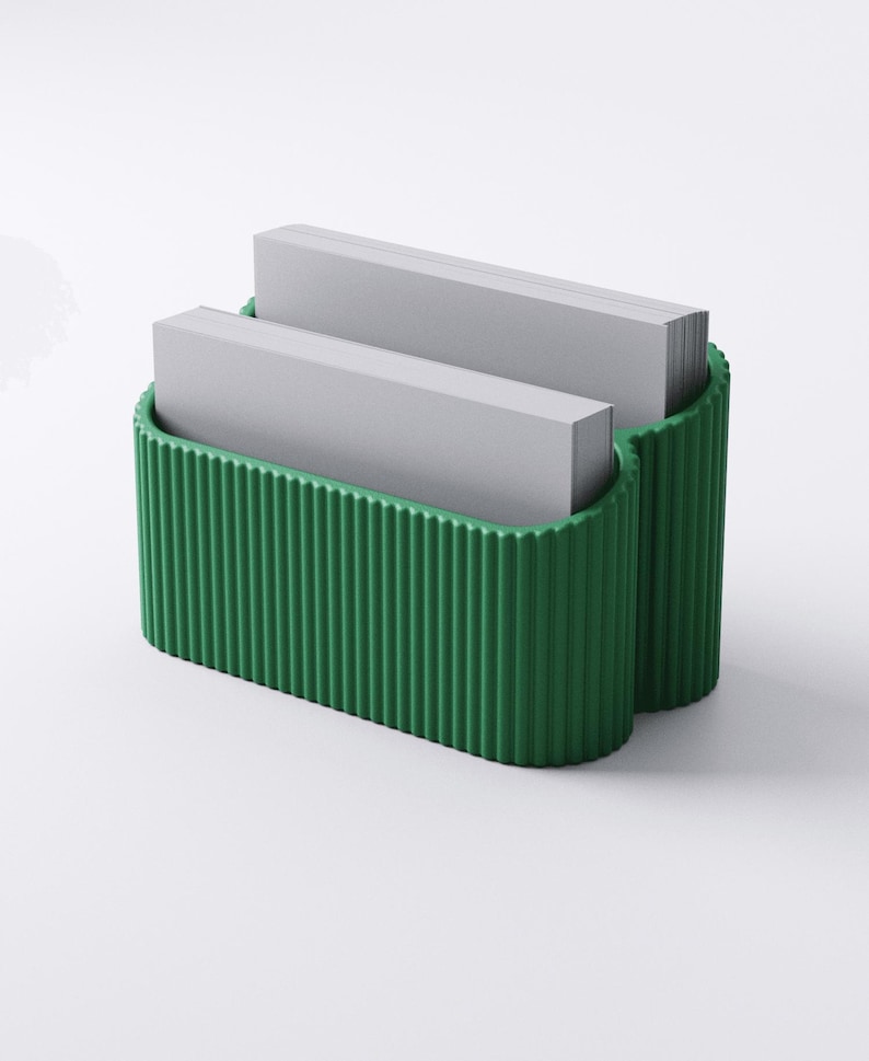 May include: A green business card holder with a ribbed exterior design. The holder is filled with two stacks of gray business cards. The holder has a curved shape and is made of a solid material.