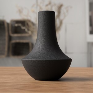 May include: A black vase with a textured surface. The vase has a wide, rounded base that tapers to a narrow neck. The vase is sitting on a wooden surface. The background is blurred, with hints of other decor.