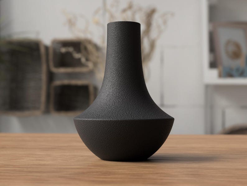 May include: A black vase with a textured surface. The vase has a wide, rounded base that tapers to a narrow neck. The vase is sitting on a wooden surface. The background is blurred, with hints of other decor.