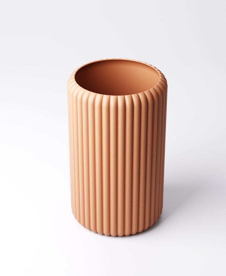 May include: A cylindrical, peach-colored vase with vertical, ridged detailing. The vase has a wide opening and a rounded base. The design is modern and minimalist, suitable for various decor styles. The vase is approximately 10 inches tall.