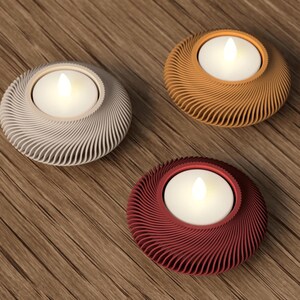 May include: Three decorative candle holders in silver, orange, and burgundy. Each holder has a circular design with a swirling pattern and holds a white LED candle. The holders are placed on a wooden surface.