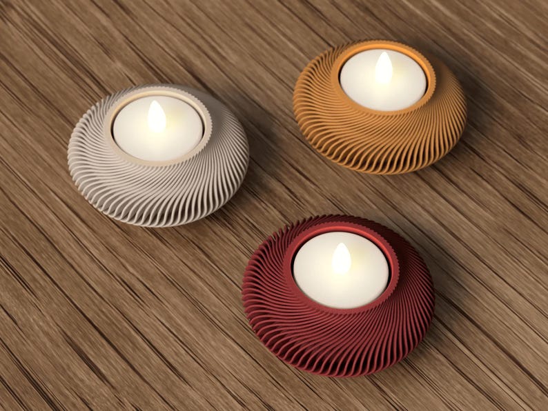 May include: Three decorative candle holders in silver, orange, and burgundy. Each holder has a circular design with a swirling pattern and holds a white LED candle. The holders are placed on a wooden surface.