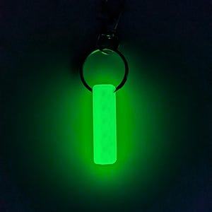May include: A glowing green keychain tag with a rectangular shape. The tag is attached to a metal keyring and a clasp. The image is set against a dark background, highlighting the bright, luminescent quality of the tag.