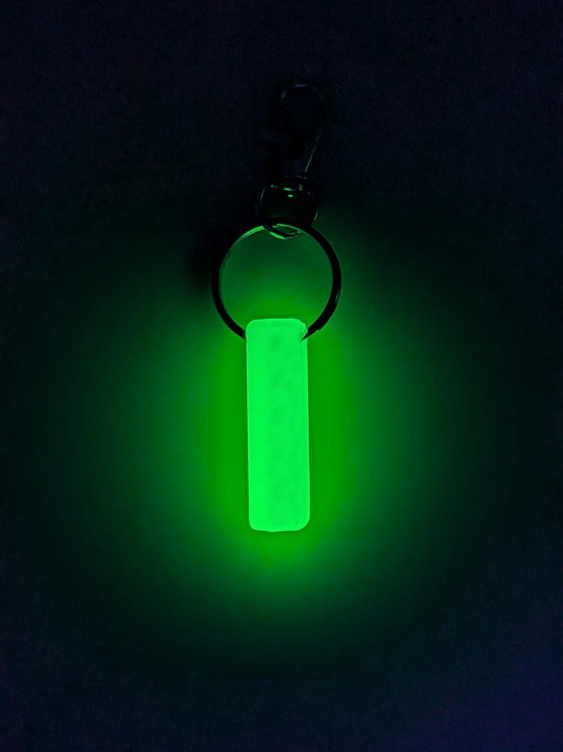 May include: A glowing green keychain tag with a rectangular shape. The tag is attached to a metal keyring and a clasp. The image is set against a dark background, highlighting the bright, luminescent quality of the tag.
