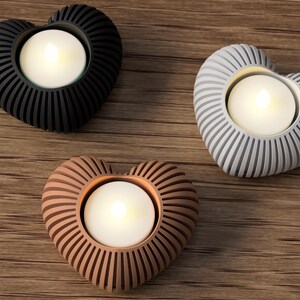 May include: Three heart-shaped candle holders in black, gray, and copper colors. Each holder has a ribbed design and holds a white, glowing tea light candle. The holders are arranged on a wooden surface.