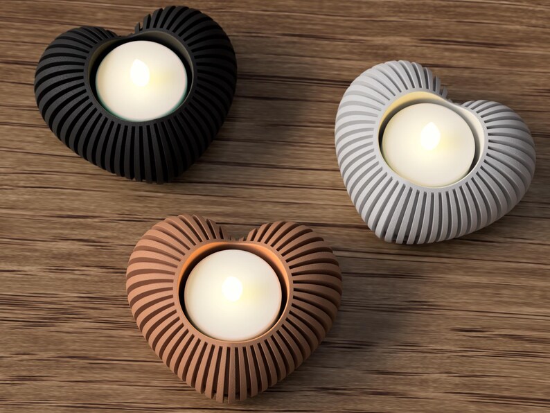 May include: Three heart-shaped candle holders in black, gray, and copper colors. Each holder has a ribbed design and holds a white, glowing tea light candle. The holders are arranged on a wooden surface.