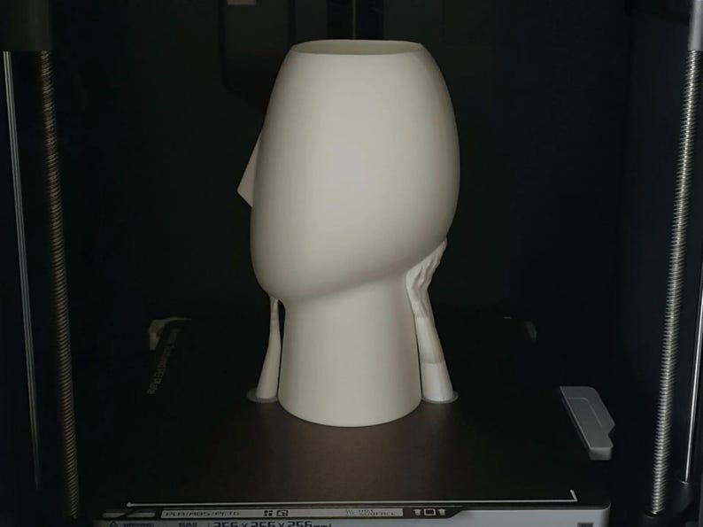 May include: A cream-colored, abstract sculpture of a head with a prominent nose and a hollow top, possibly a vase or planter. The sculpture has a smooth surface and is set against a dark background.