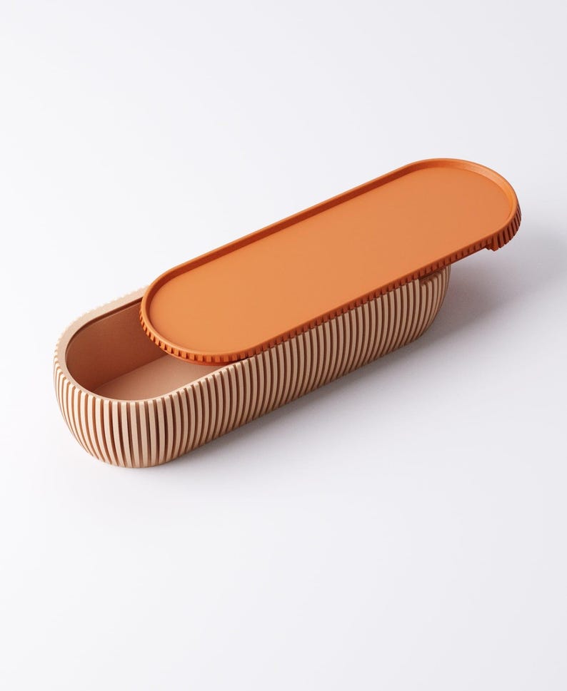 May include: An oval-shaped wooden box with a lid. The box is made of vertical wooden slats, and the lid is a solid orange color. The interior of the box is a light peach color. The lid is slightly ajar, revealing the interior.