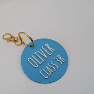 May include: A light blue circular keychain tag with the name "OLIVER" and "CLASS 3B" in white raised lettering. The tag is attached to a gold-colored metal clasp and ring.