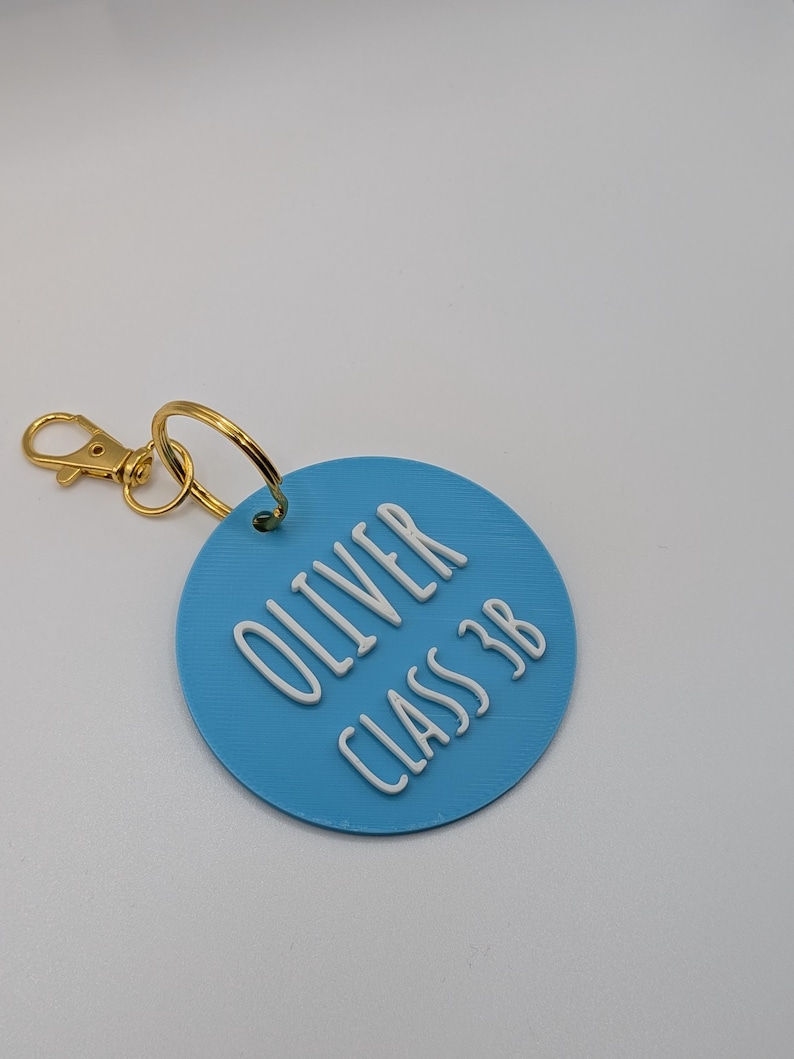 May include: A light blue circular keychain tag with the name "OLIVER" and "CLASS 3B" in white raised lettering. The tag is attached to a gold-colored metal clasp and ring.