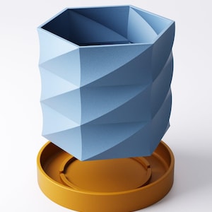 May include: A geometric blue vase with a textured surface, resting on a round, mustard-yellow base. The vase has a hexagonal top and a spiraling, faceted design. The base has a circular indentation.