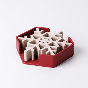 May include: A set of snowflake-shaped coasters in a red holder. The coasters are a light beige color and are stacked neatly inside the red, hexagonal-shaped holder. The coasters are designed to resemble snowflakes.