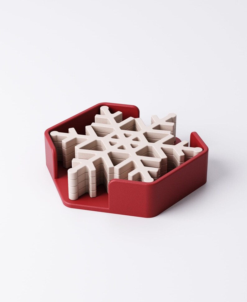 May include: A set of snowflake-shaped coasters in a red holder. The coasters are a light beige color and are stacked neatly inside the red, hexagonal-shaped holder. The coasters are designed to resemble snowflakes.