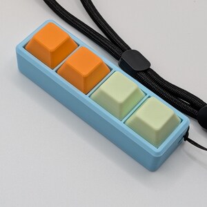 May include: A light blue rectangular device with four square buttons in orange and light green. The device is attached to a black lanyard. The buttons are arranged in two rows of two.