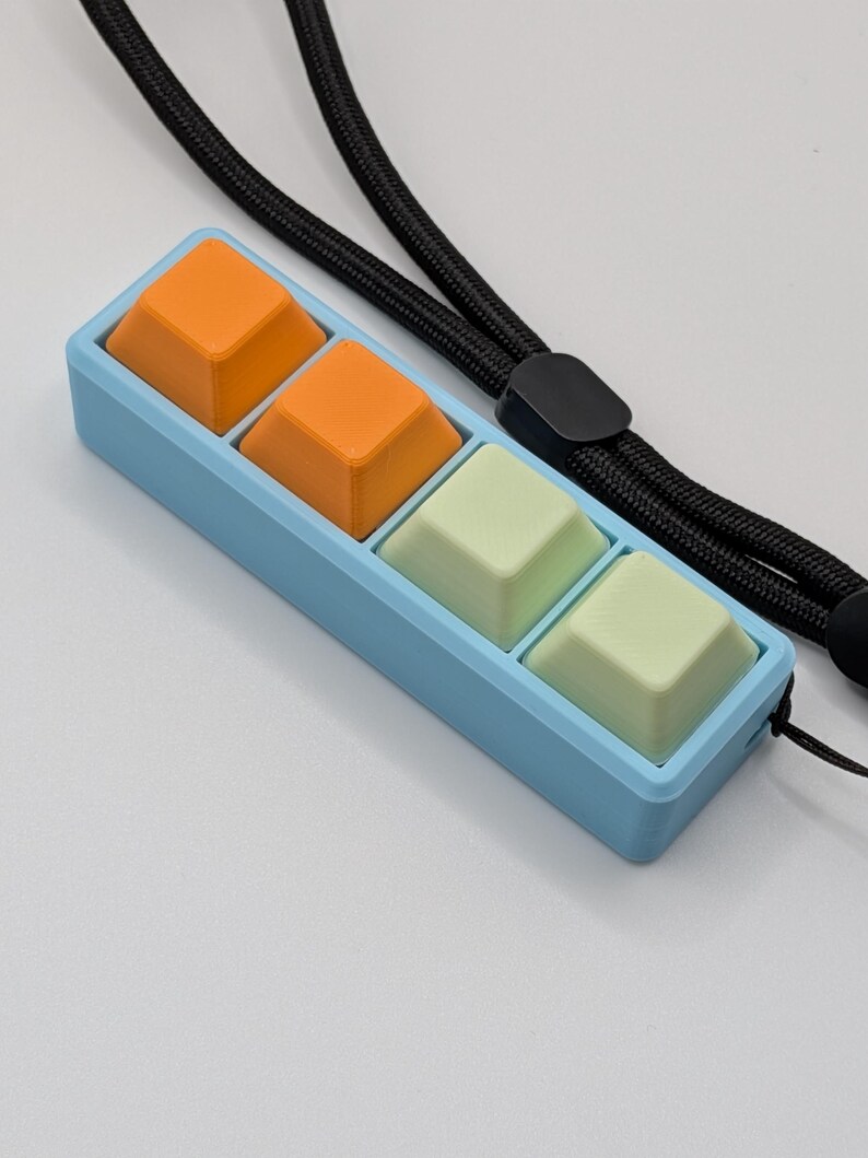 May include: A light blue rectangular device with four square buttons in orange and light green. The device is attached to a black lanyard. The buttons are arranged in two rows of two.