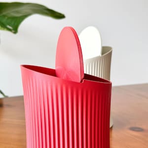 May include: A bright red waste bin with a vertical ribbed texture and a matching red swing lid. Positioned behind it is a second waste bin in a neutral off-white colour, also featuring a vertical ribbed texture and a matching swing lid.