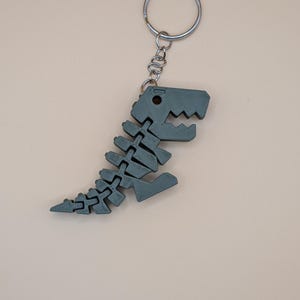 May include: A gray dinosaur skeleton keychain. The keychain features a detailed, articulated design of a Tyrannosaurus Rex skeleton. The dinosaur is attached to a silver metal keyring and chain, perfect for keys or as a decorative accessory.