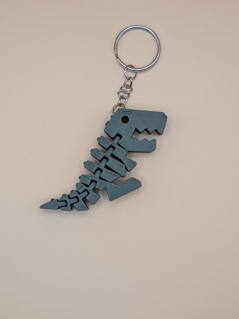 May include: A gray dinosaur skeleton keychain. The keychain features a detailed, articulated design of a Tyrannosaurus Rex skeleton. The dinosaur is attached to a silver metal keyring and chain, perfect for keys or as a decorative accessory.