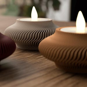 May include: Three decorative candle holders with a wavy, textured design. The candle holders are in shades of burgundy, gray, and brown, each holding a glowing LED candle. The design is modern and minimalist, suitable for home decor.