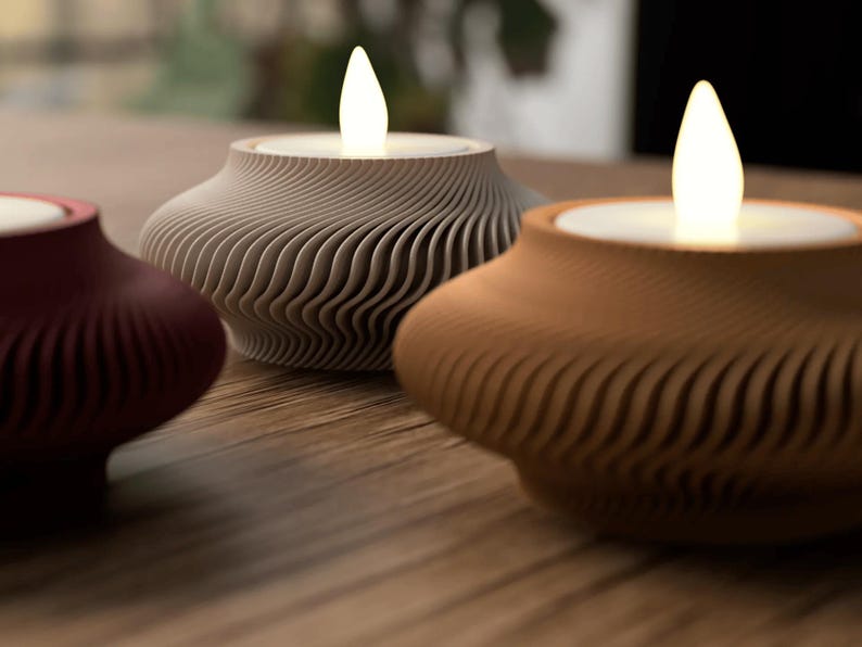 May include: Three decorative candle holders with a wavy, textured design. The candle holders are in shades of burgundy, gray, and brown, each holding a glowing LED candle. The design is modern and minimalist, suitable for home decor.