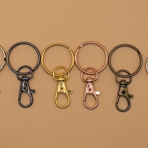 May include: A collection of keychains in various metallic finishes, including silver, black, gold, rose gold, and bronze. Each keychain features a round ring and a lobster clasp for attaching keys or other items. The keychains are arranged on a neutral background.