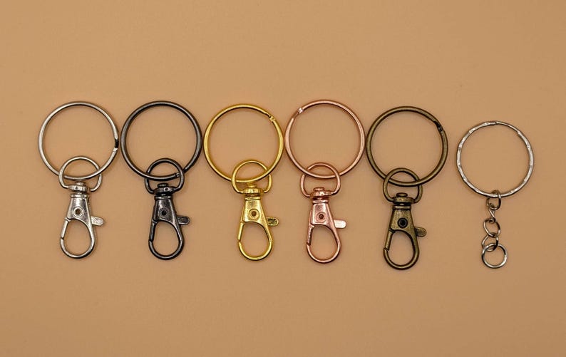 May include: A collection of keychains in various metallic finishes, including silver, black, gold, rose gold, and bronze. Each keychain features a round ring and a lobster clasp for attaching keys or other items. The keychains are arranged on a neutral background.