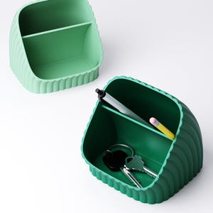 May include: Two green desk tidies with a ribbed design. One is a lighter shade of green and empty, while the other is a darker green and contains a black pen, a yellow pencil, and a set of keys. The tidies have a divided interior.