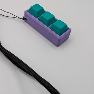 May include: A custom-made, 3D-printed keycap holder with three teal keycaps set in a purple base. A black lanyard is attached for portability. The keycap holder is designed for a specific keyboard or project.